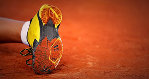 Ein gelber Sportschuh mit schwarzer Sohle liegt auf einem roten Sandplatz.