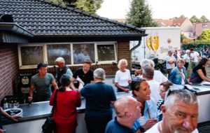 Eine Gruppe von Menschen steht drau&szlig;en vor einem Geb&auml;ude mit einem dunklen Dach, einige bedienen sich an einem Tresen. Im Hintergrund sind weitere Personen und B&auml;ume zu sehen.