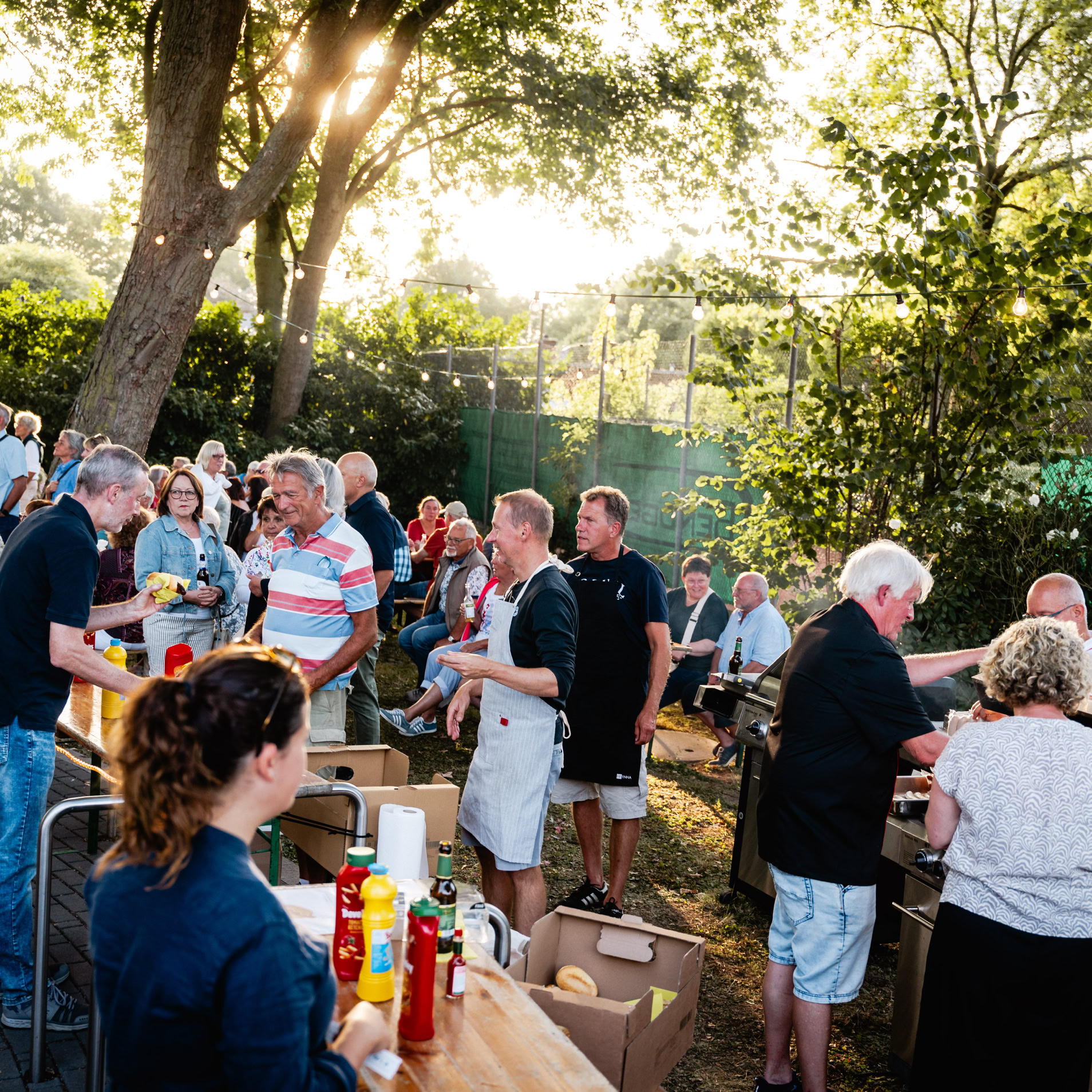 Gruppe von Menschen bei einer Outdoor-Veranstaltung mit Tischen und Getr&auml;nken, umgeben von B&auml;umen und Sonnenlicht.