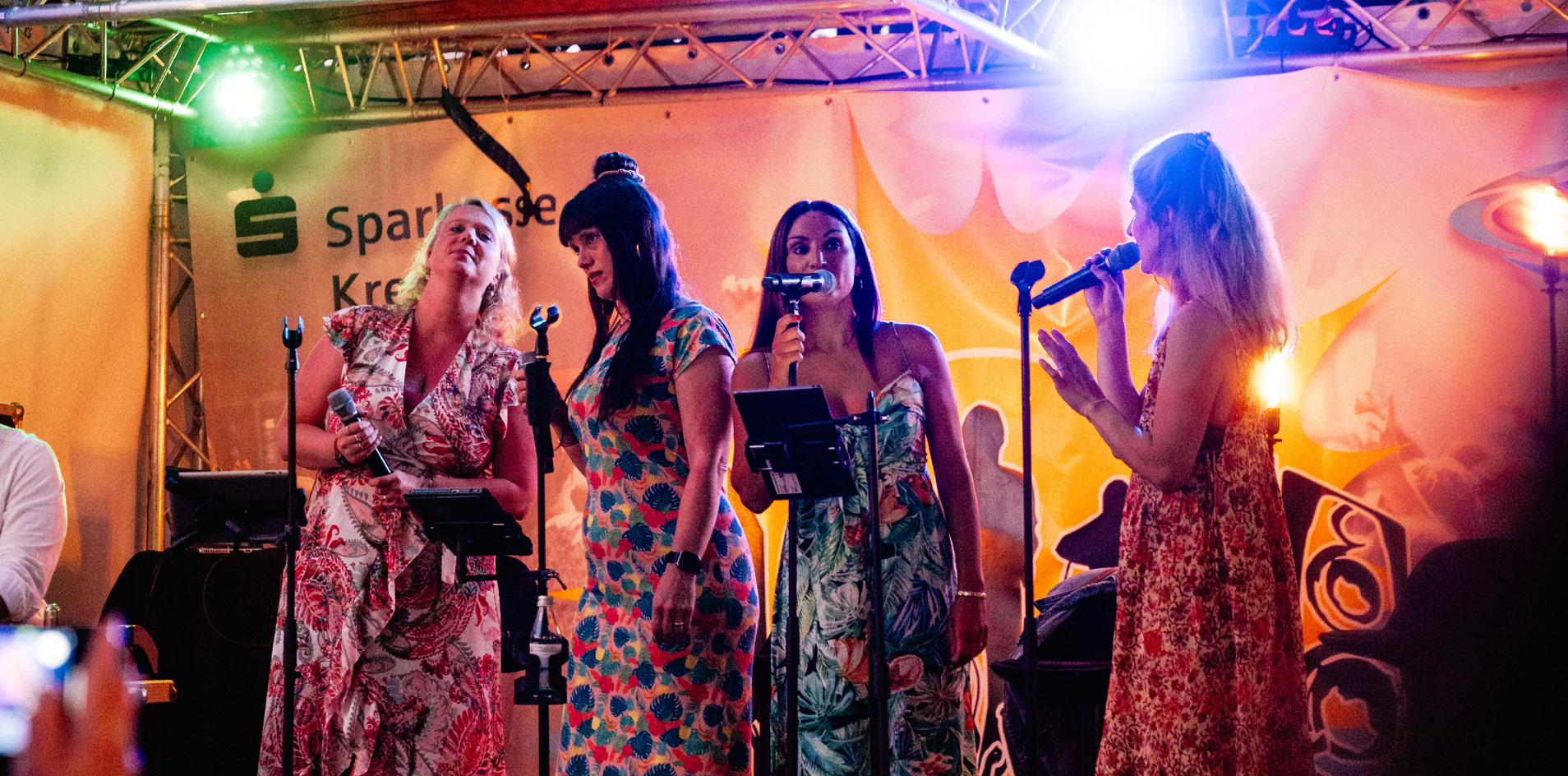 Vier Personen singen auf einer B&uuml;hne unter bunten Lichtern, umgeben von Pflanzen.