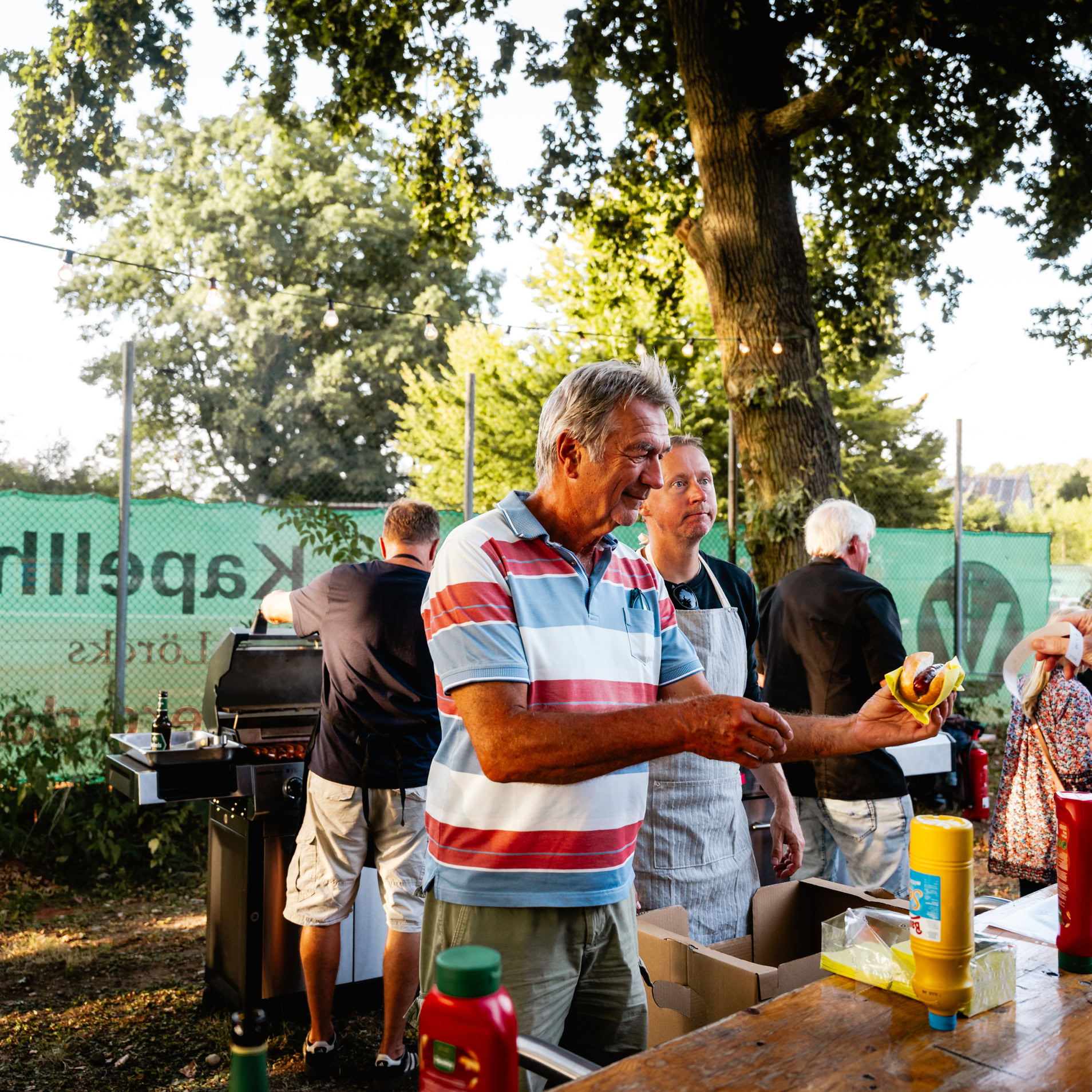Ein Mann in einem gestreiften Hemd erh&auml;lt Essen von einem anderen Mann an einem Stand im Freien, umgeben von B&auml;umen und einem gr&uuml;nen Banner im Hintergrund.