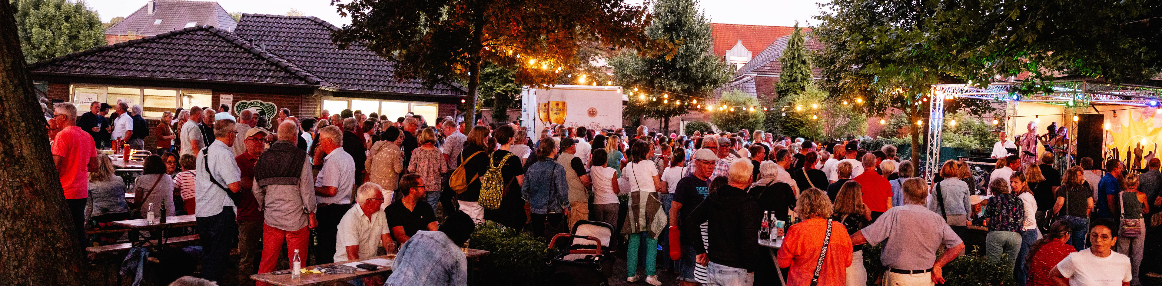 Eine Menschenmenge versammelt sich bei einem Freiluftfest mit B&auml;umen und Geb&auml;uden im Hintergrund.
