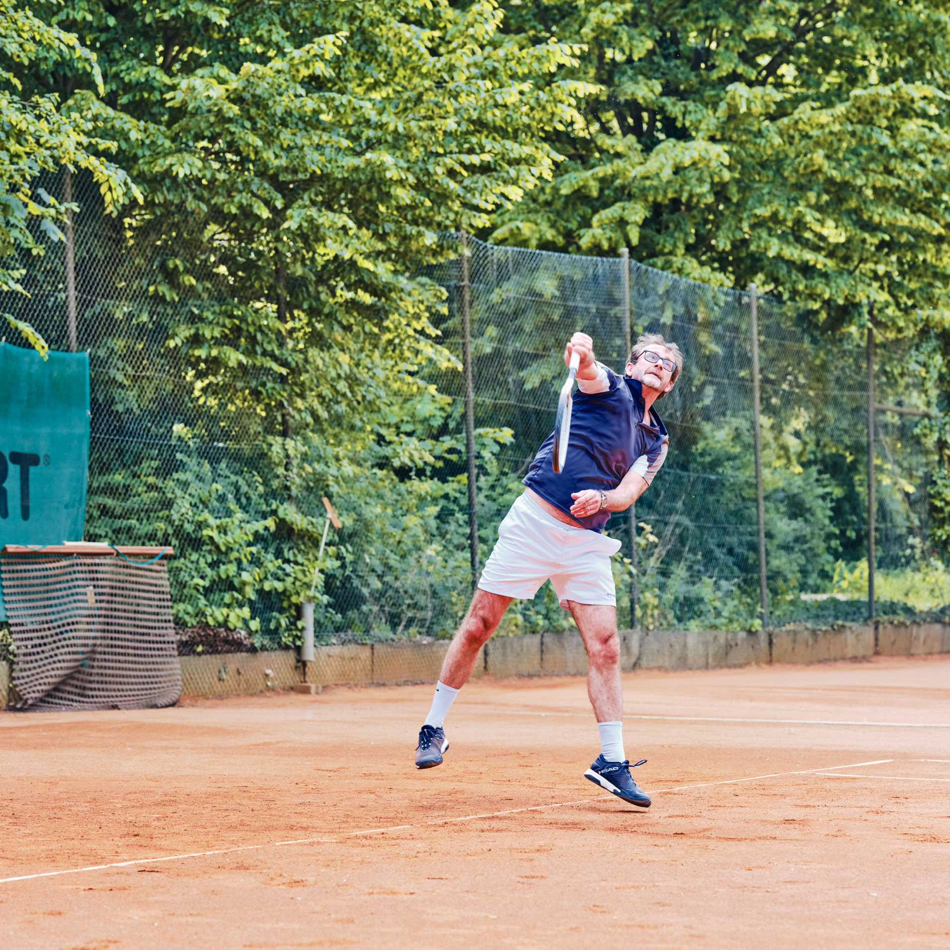 Person spielt Tennis auf einem Sandplatz, umgeben von B&auml;umen.