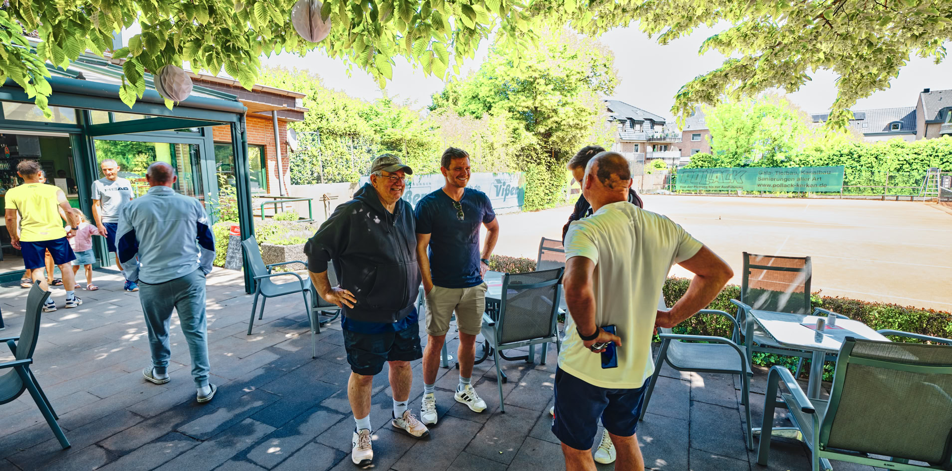 Mehrere Personen stehen auf einer Terrasse unter B&auml;umen, umgeben von Tischen und St&uuml;hlen. Im Hintergrund ist ein Geb&auml;ude und ein Tennisplatz zu sehen.