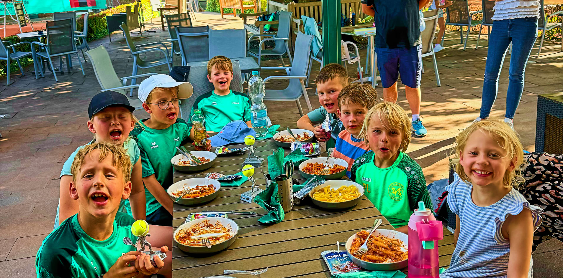 Kinder sitzen an einem Tisch im Freien und essen. Im Hintergrund stehen zwei Erwachsene. Der Bereich ist von B&auml;umen umgeben.