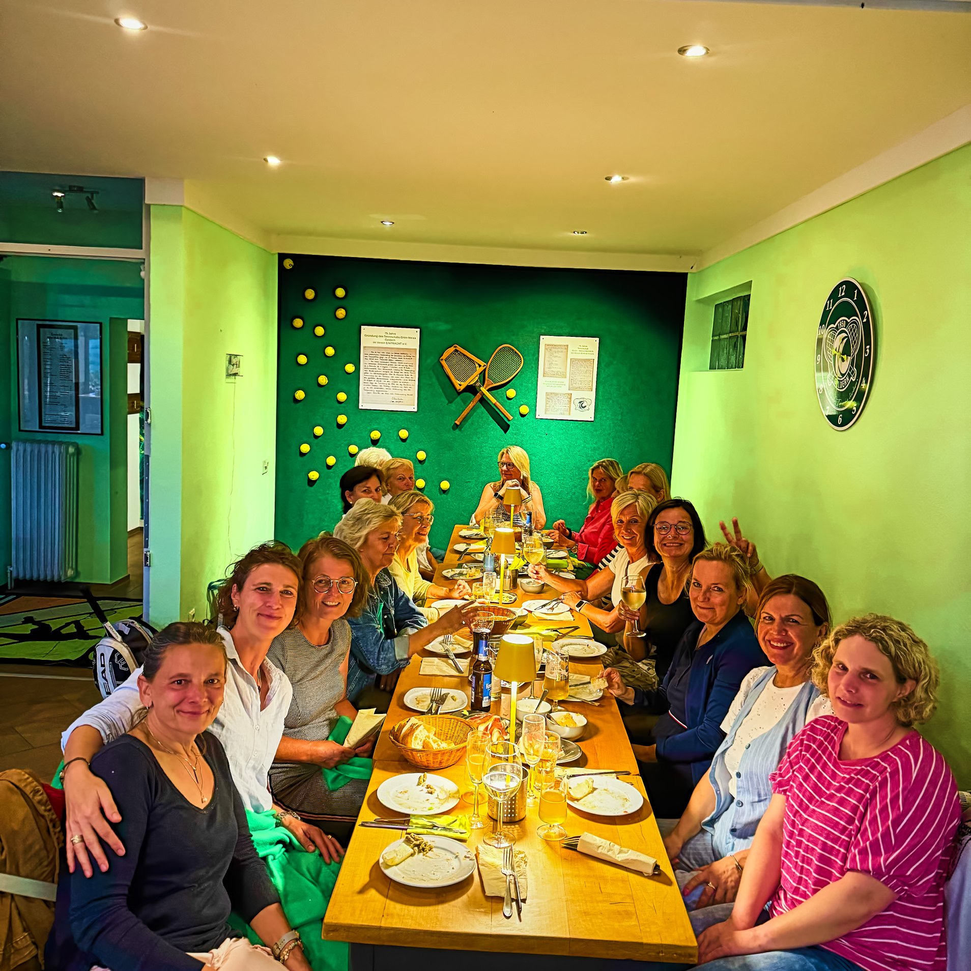 Eine Gruppe von Menschen sitzt an einem langen Tisch in einem Raum mit gr&uuml;nen W&auml;nden. Auf dem Tisch stehen Teller und Gl&auml;ser.