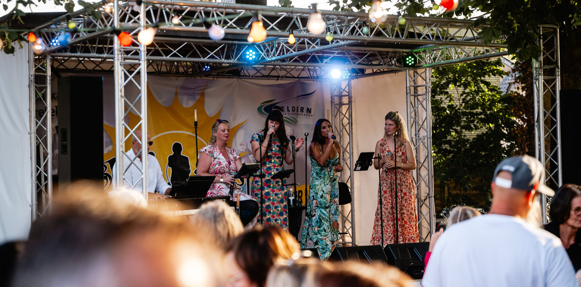 Eine Gruppe von Menschen steht auf einer B&uuml;hne im Freien, umgeben von B&auml;umen und Lichtern, w&auml;hrend eine Menschenmenge im Vordergrund zuschaut.