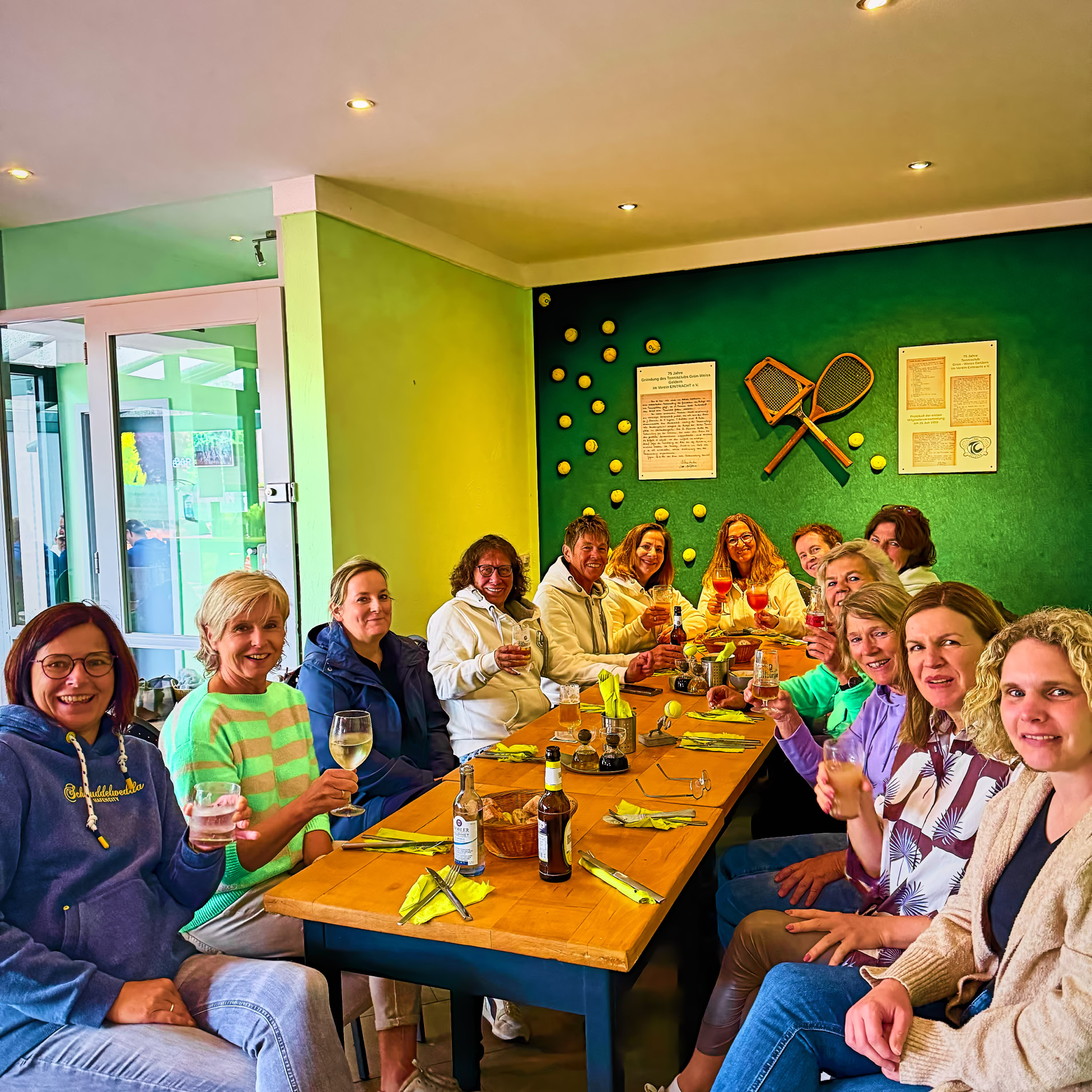 Eine Gruppe von Menschen sitzt an einem langen Tisch in einem Raum mit gr&uuml;nen W&auml;nden, dekoriert mit Tennisschl&auml;gern und B&auml;llen.