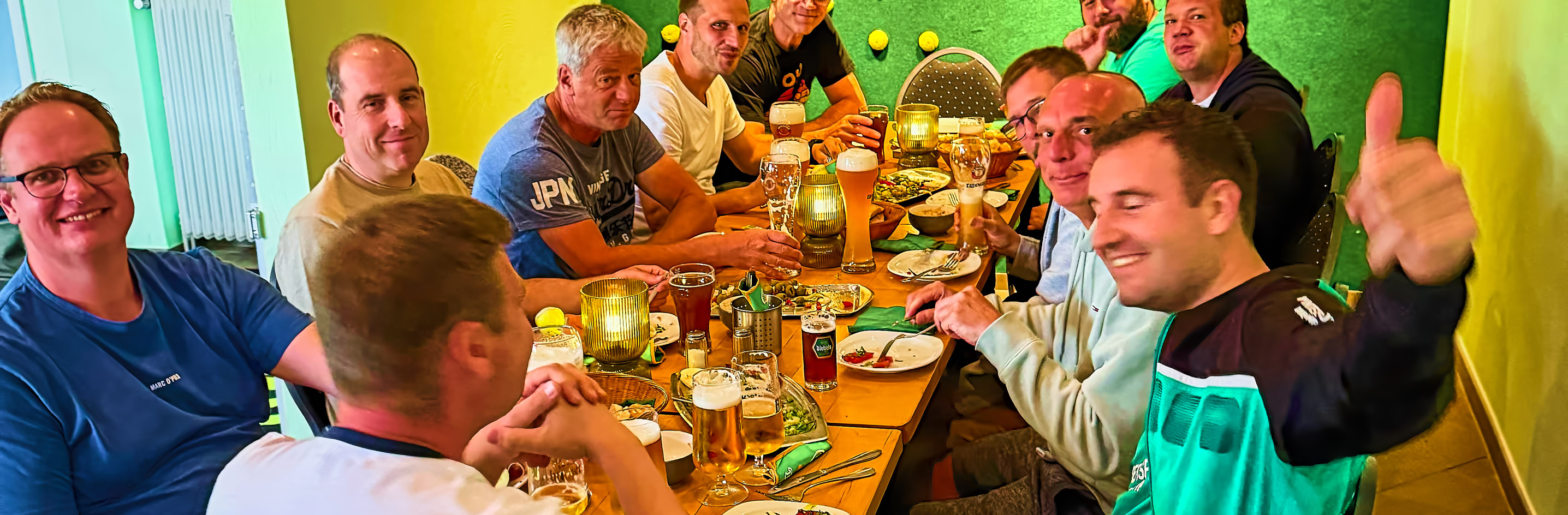 Eine Gruppe von neun Personen sitzt um einen Tisch in einem Restaurant mit gr&uuml;ner Wand, auf der Tennisschl&auml;ger und B&auml;lle dekoriert sind.