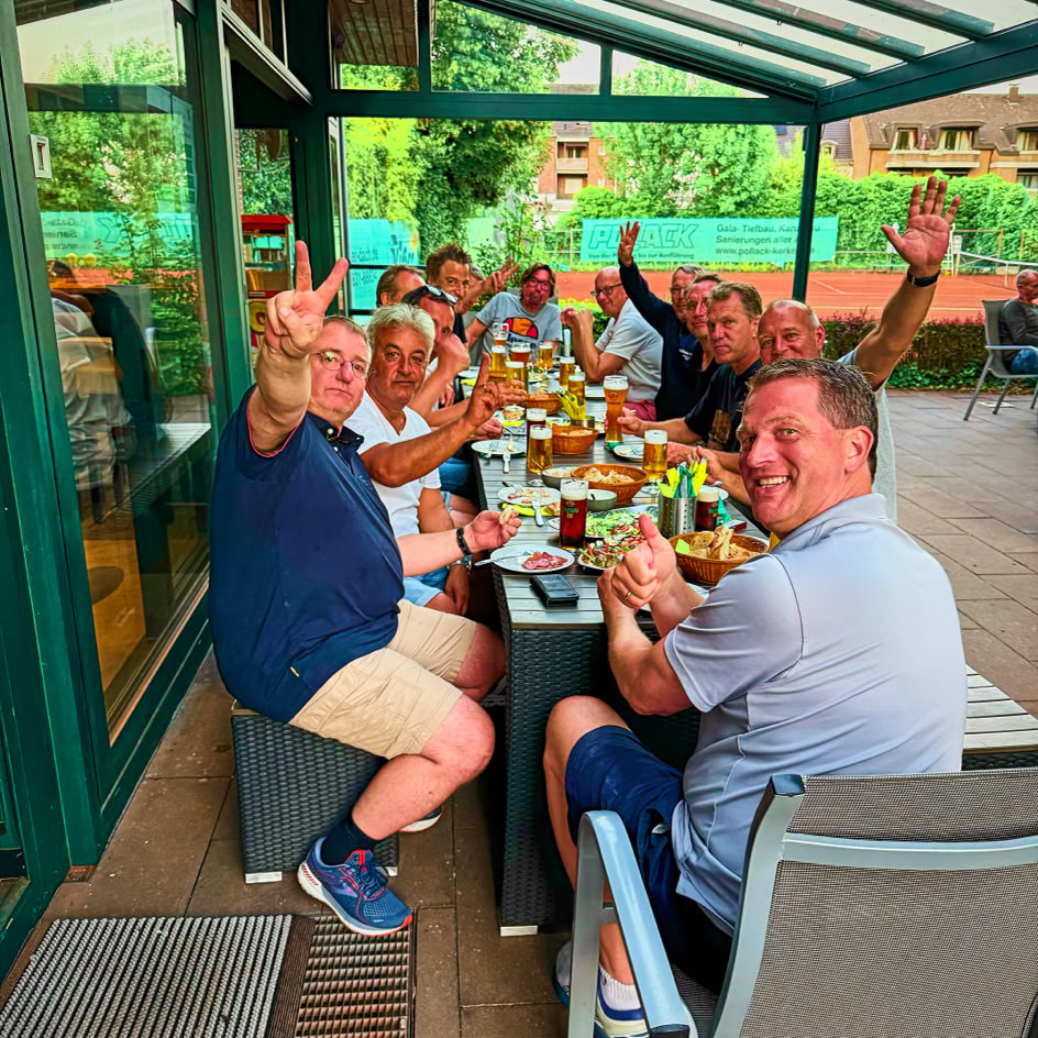 Eine Gruppe von Menschen sitzt an einem langen Tisch im Freien und hebt die H&auml;nde, w&auml;hrend sie Mahlzeiten und Getr&auml;nke genie&szlig;en.