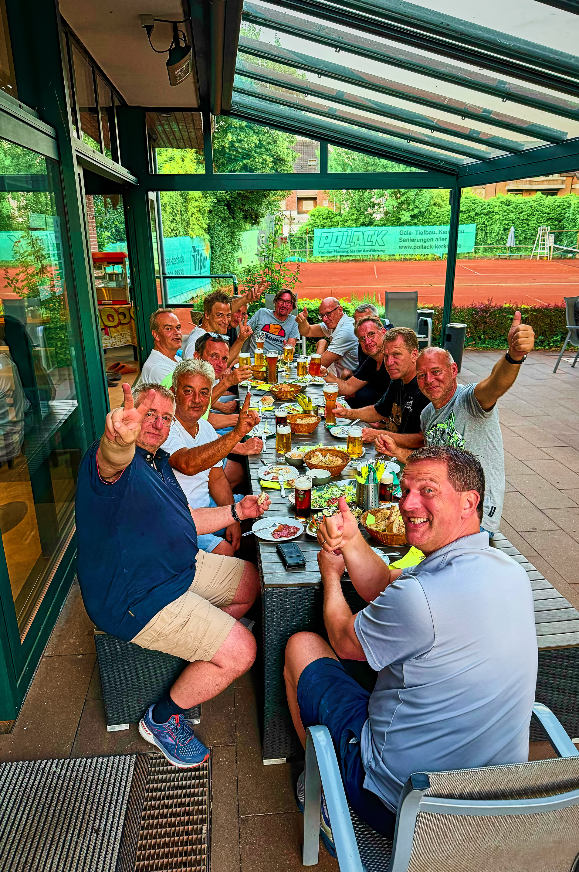 Gruppe von Menschen sitzt an einem langen Tisch im Freien, isst und hebt Gl&auml;ser zum Ansto&szlig;en.