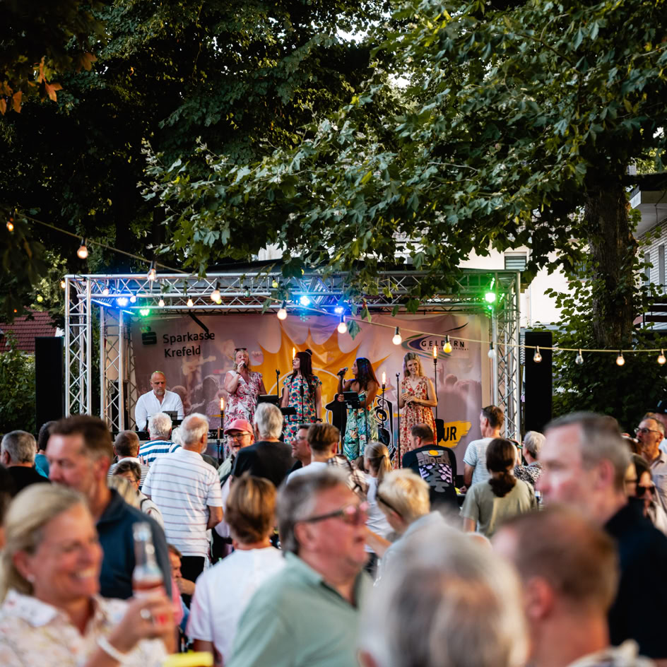 Menschenmenge bei einer Veranstaltung im Freien mit einer B&uuml;hne im Hintergrund, auf der Musiker auftreten; umgeben von B&auml;umen und Lichtern.