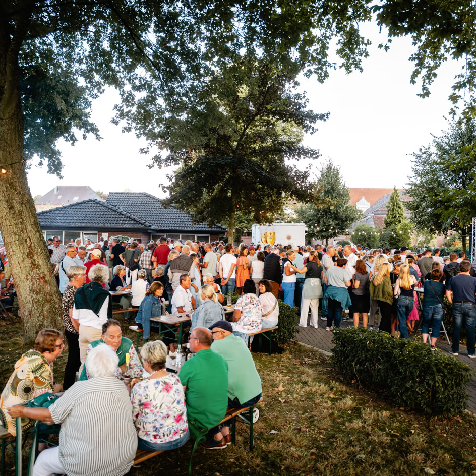 Menschenmenge bei einem Freiluftfest mit B&auml;umen im Hintergrund, einige sitzen an Tischen, andere stehen.