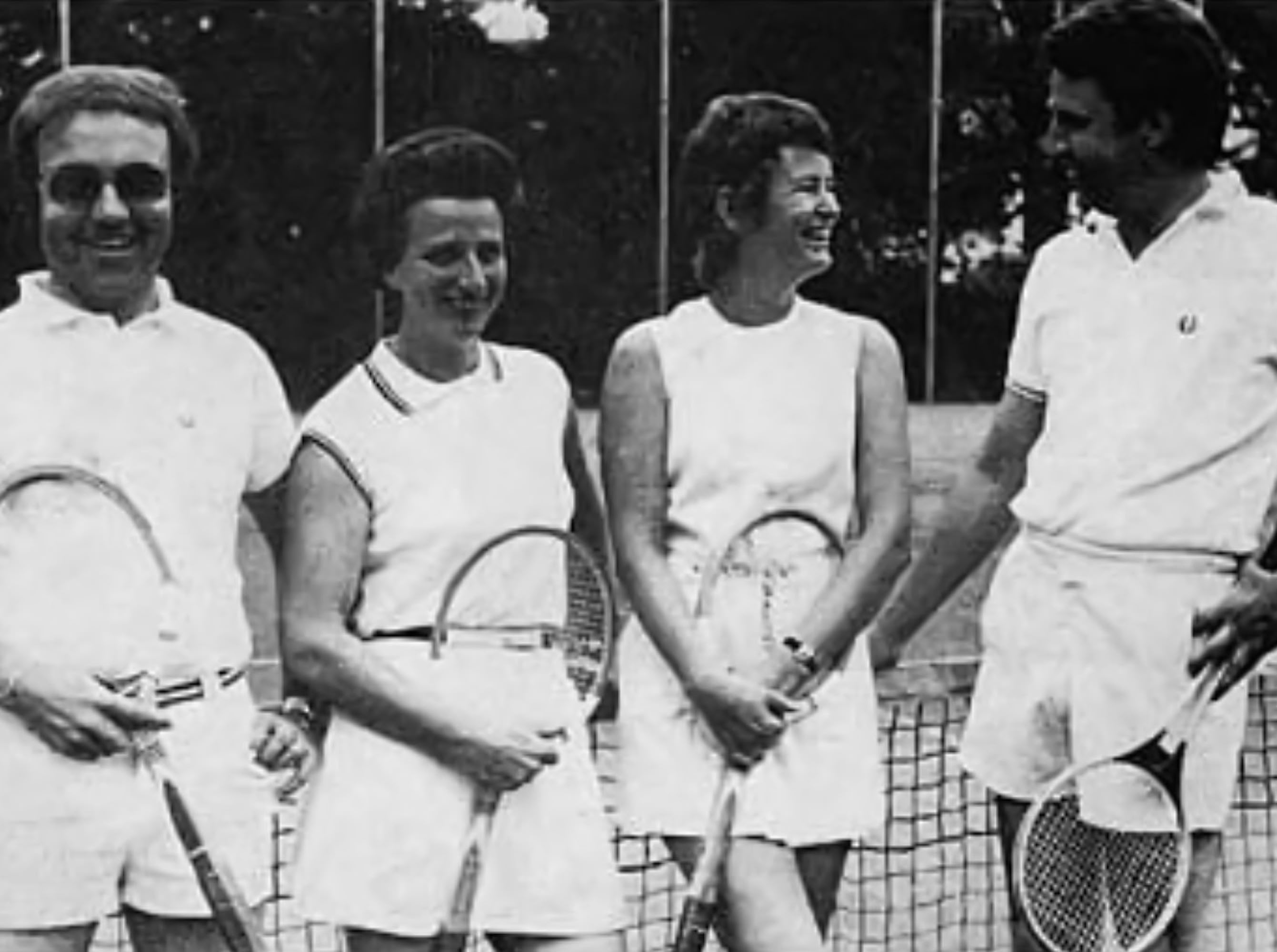 Vier Personen in Tennisbekleidung stehen auf einem Tennisplatz und halten Tennisschl&auml;ger.