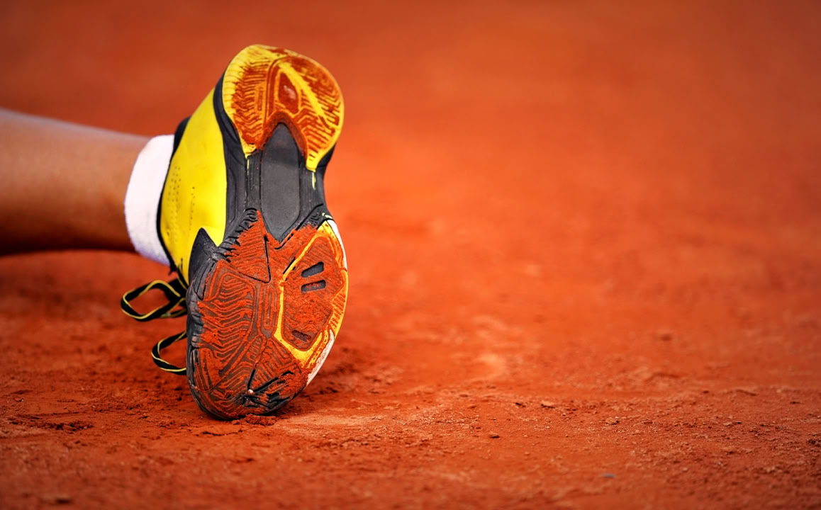 Gelber Sportschuh auf rotem Sandboden, Bein eines Sportlers sichtbar.