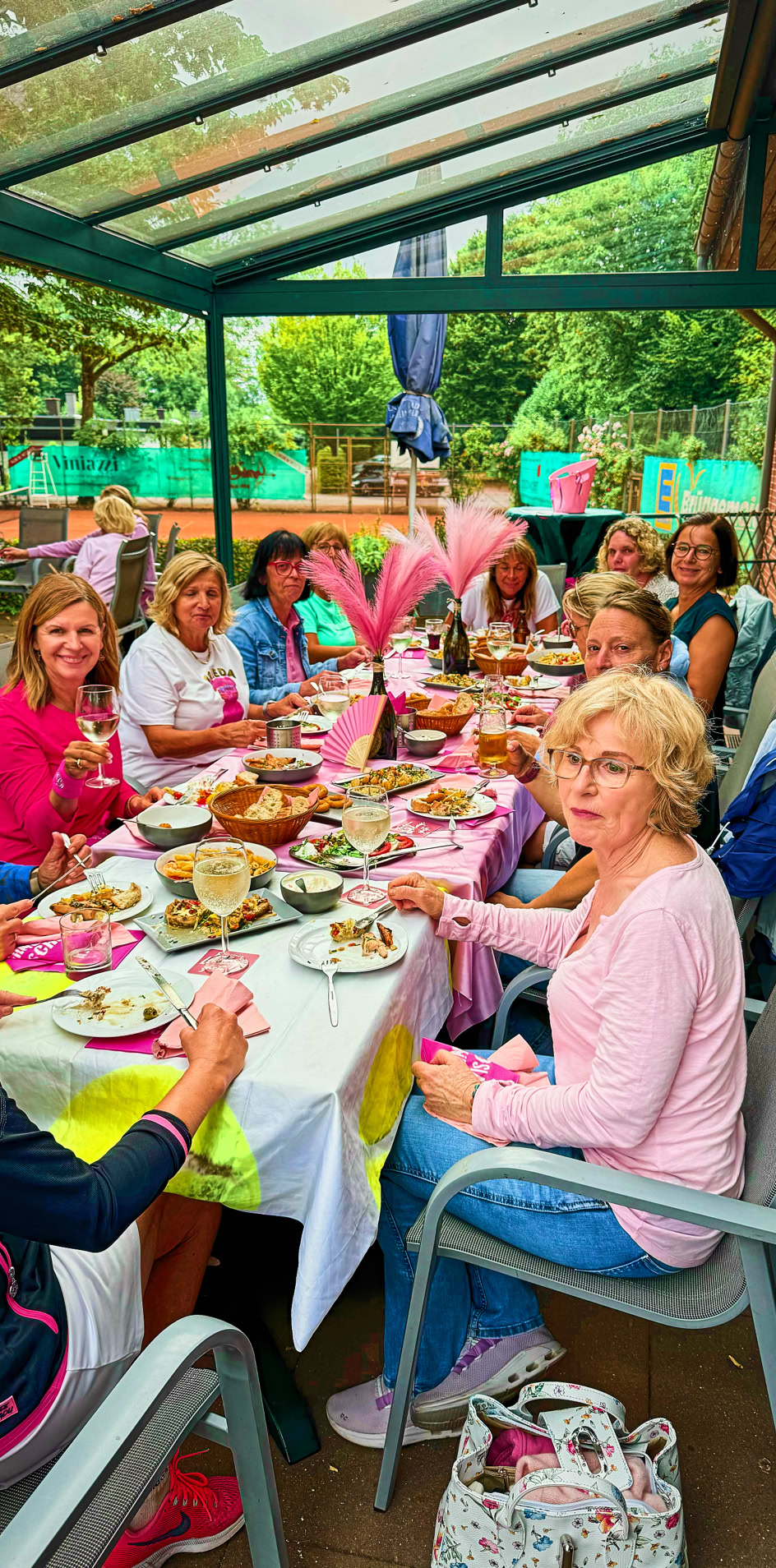 Eine Gruppe von Frauen sitzt an einem gedeckten Tisch unter einem Glasdach, umgeben von gr&uuml;ner Vegetation.