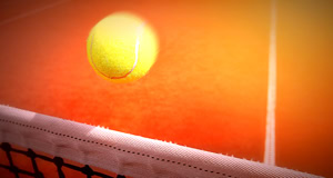 Ein gelber Tennisball fliegt &uuml;ber ein Tennisnetz auf einem Sandplatz.