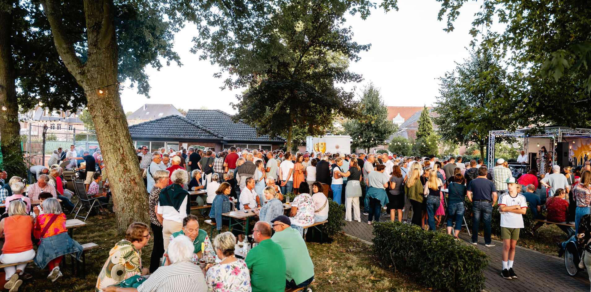 Eine gro&szlig;e Menschenmenge versammelt sich im Freien unter B&auml;umen, einige sitzen an Tischen, w&auml;hrend andere stehen. Im Hintergrund sind Geb&auml;ude und ein klarer Himmel zu sehen.