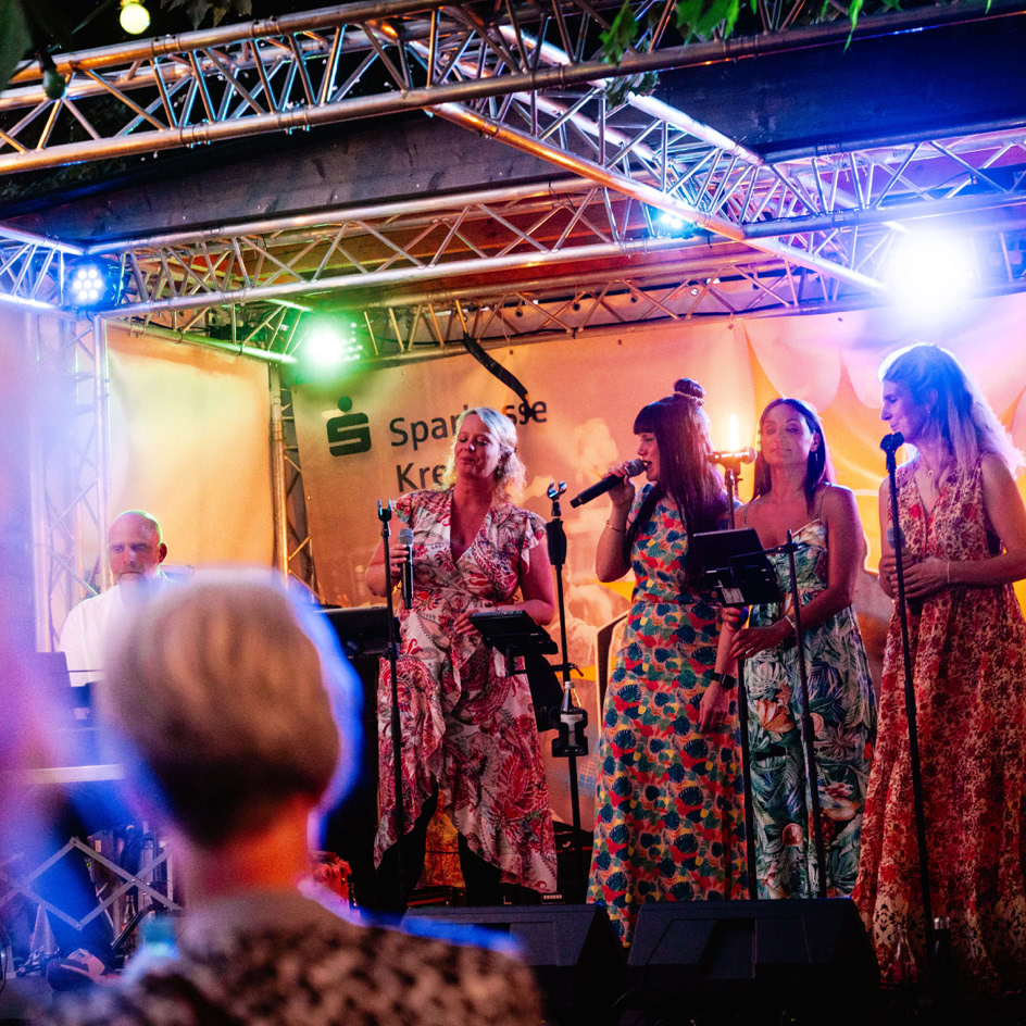 Eine Gruppe von Frauen in bunten Kleidern singt auf einer B&uuml;hne mit bunten Lichtern, w&auml;hrend ein Mann am Keyboard spielt. Im Vordergrund ist der Hinterkopf einer Person zu sehen.