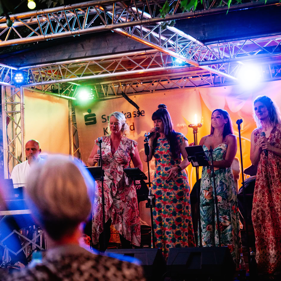 Eine Gruppe von Menschen steht auf einer B&uuml;hne mit bunten Lichtern und Mikrofonen, w&auml;hrend im Vordergrund unscharf K&ouml;pfe von Zuschauern zu sehen sind.