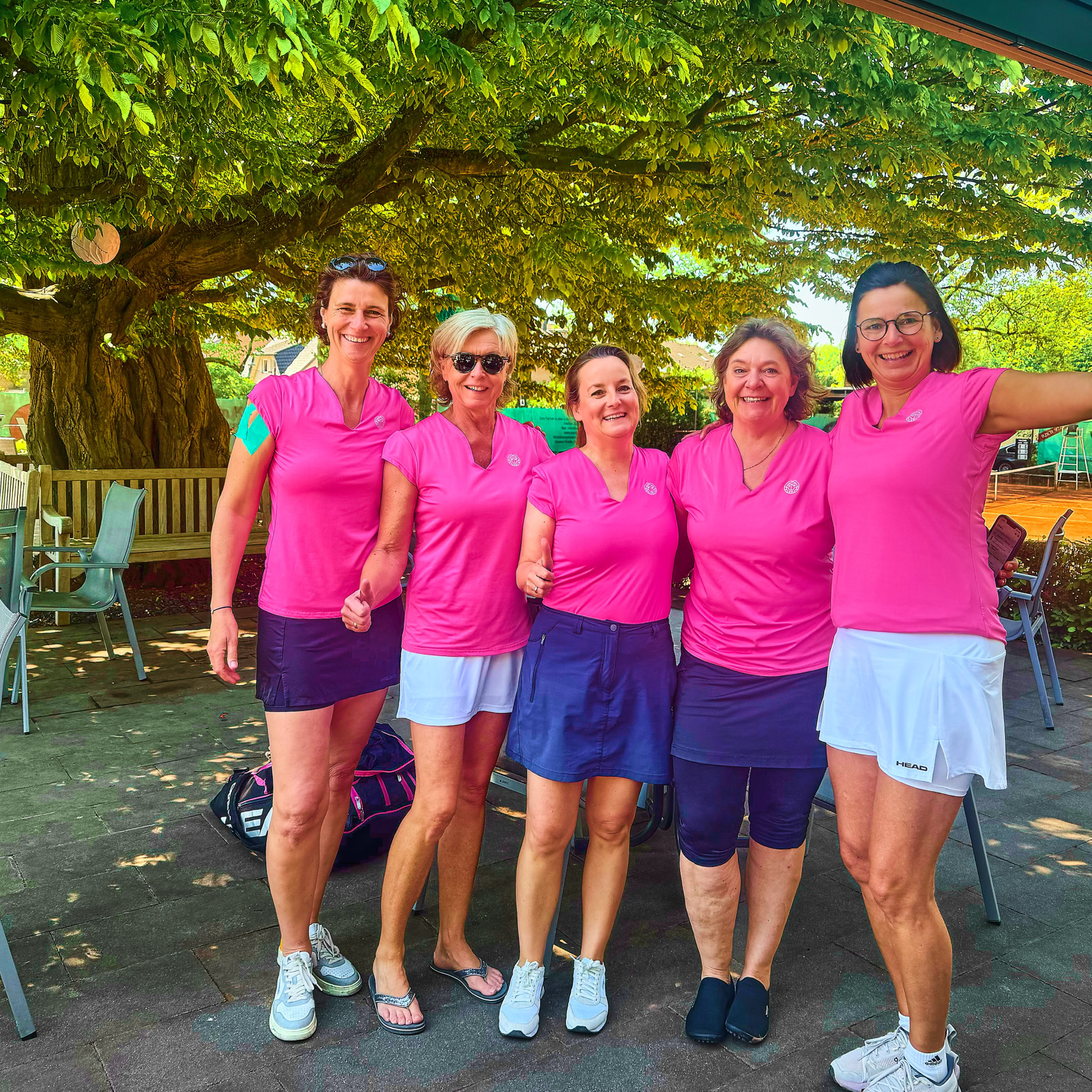 F&uuml;nf Personen in pinken Oberteilen und Sportkleidung stehen l&auml;chelnd nebeneinander im Freien, umgeben von B&auml;umen und St&uuml;hlen.