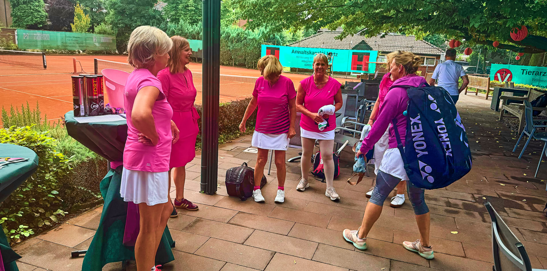 Eine Gruppe von Frauen in pinker Sportkleidung steht auf einer Terrasse neben einem Tennisplatz.