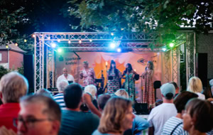 Eine Gruppe von Menschen steht vor einer B&uuml;hne im Freien, auf der mehrere Personen unter bunten Lichtern auftreten.