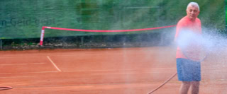 Eine Person bew&auml;ssert einen Tennisplatz mit einem Schlauch.