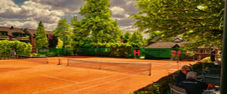 Ein Tennisplatz mit rotem Sand, umgeben von B&auml;umen und B&uuml;schen, mit mehreren Personen, die auf und neben dem Platz stehen.