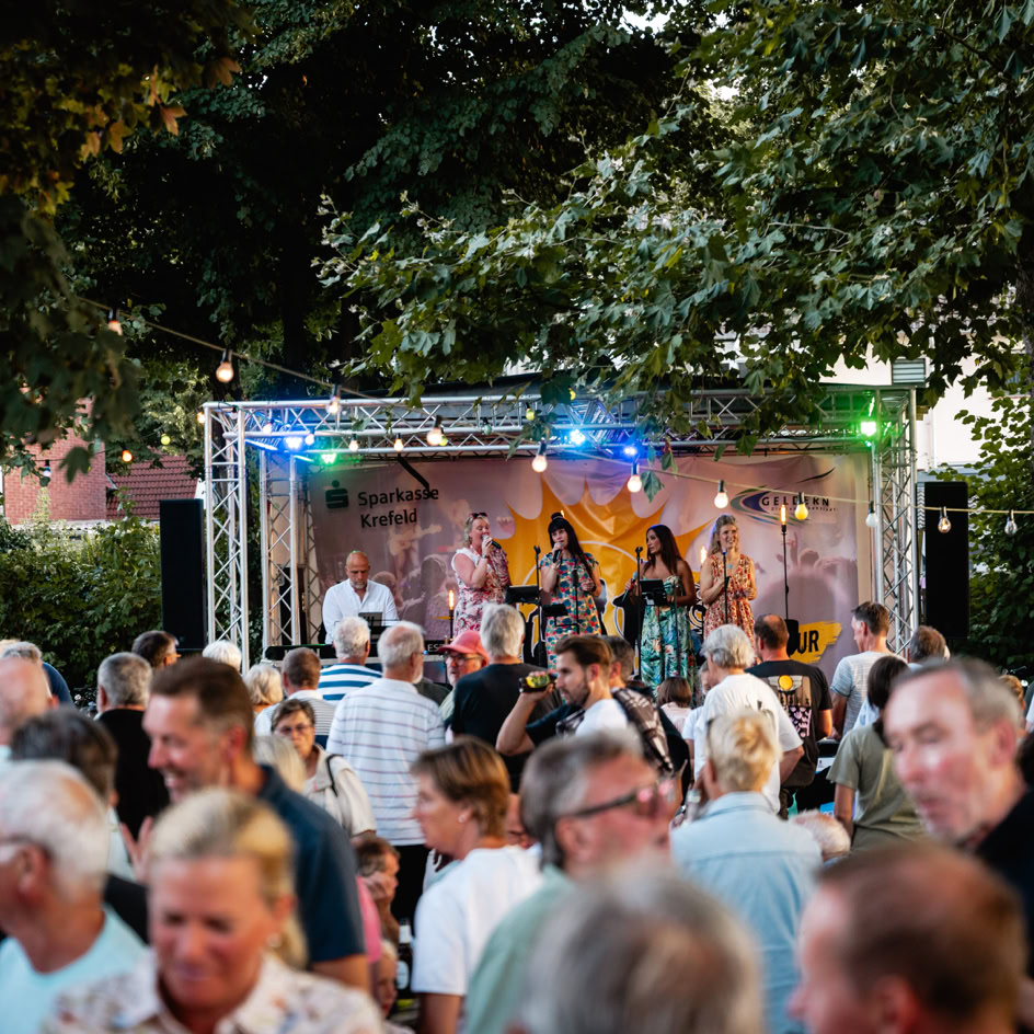 Menschenmenge vor einer B&uuml;hne im Freien, auf der eine Band spielt, umgeben von B&auml;umen und Lichtern.