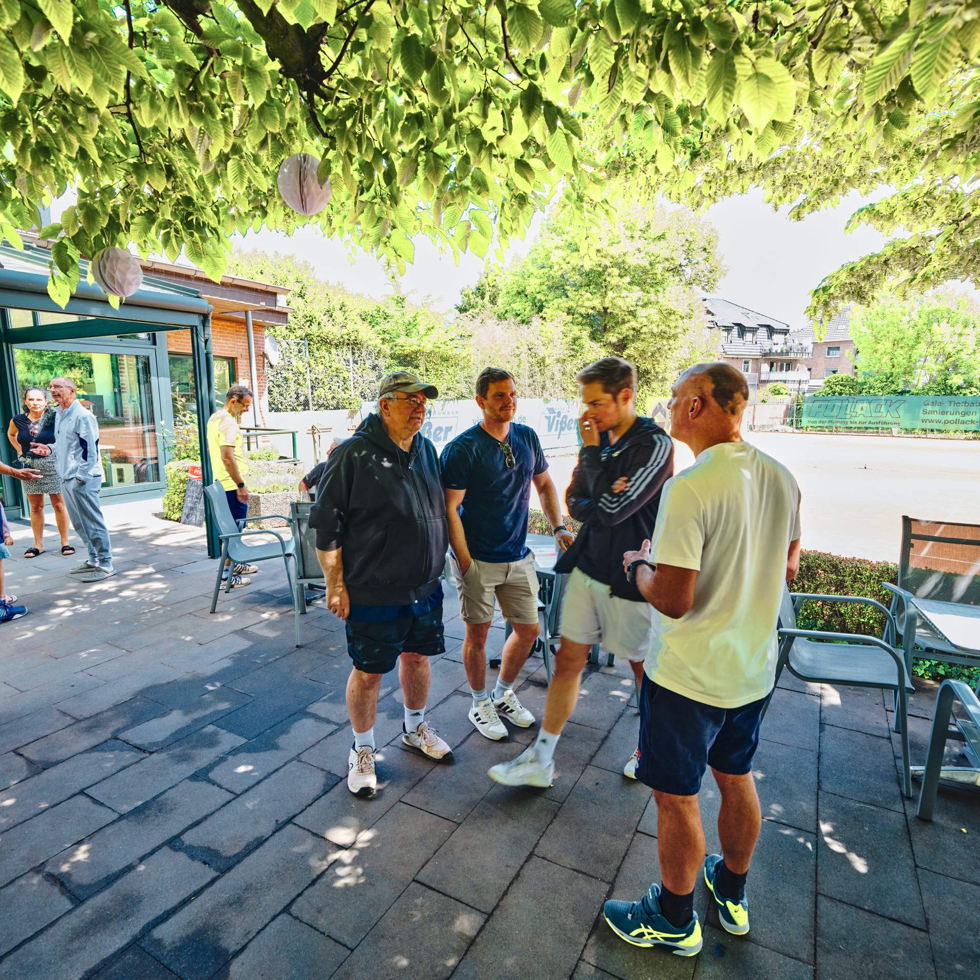 Mehrere Personen stehen und unterhalten sich auf einer Terrasse mit Tischen und St&uuml;hlen, umgeben von B&auml;umen und Pflanzen.