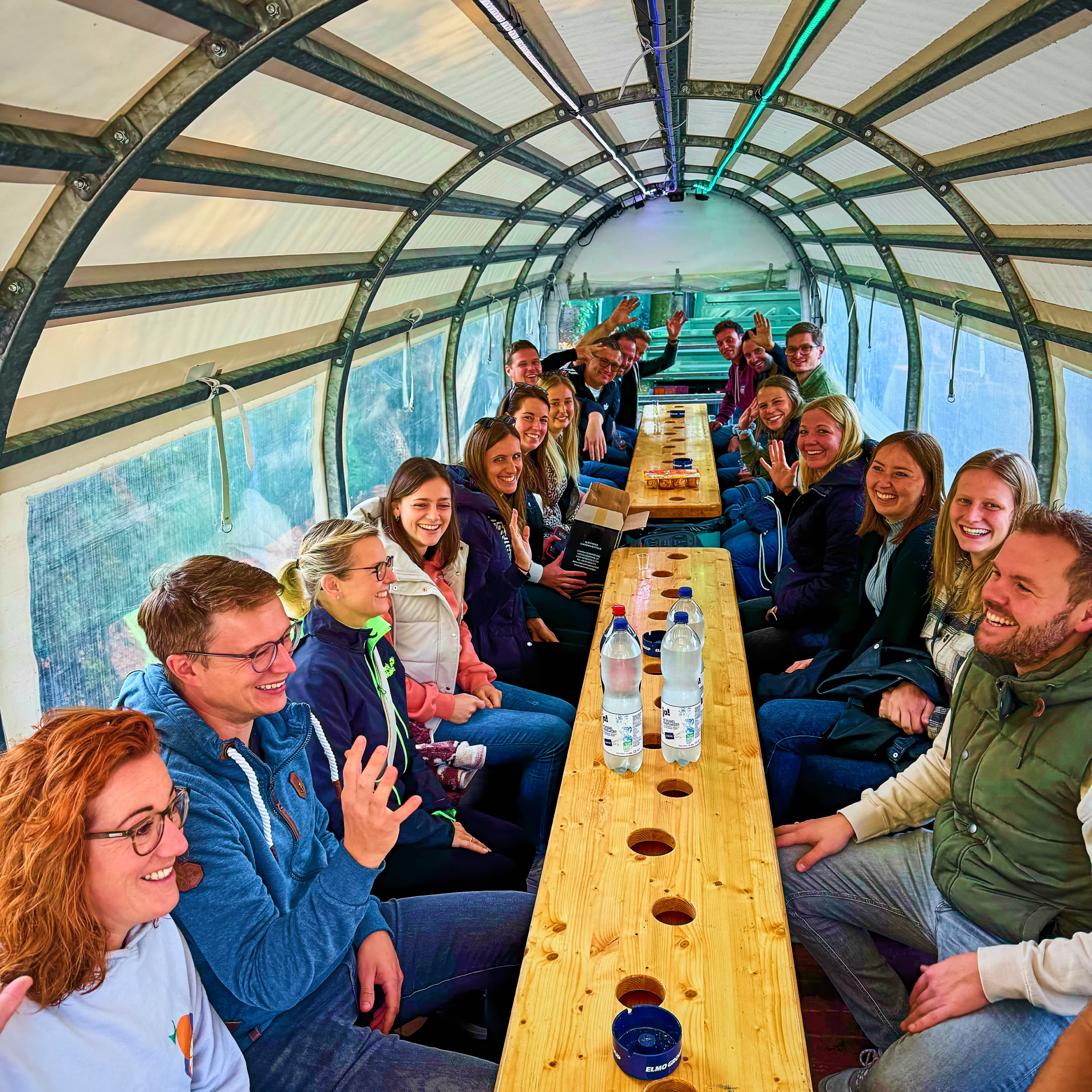 Eine Gruppe von Menschen sitzt an einem langen Holztisch in einem &uuml;berdachten Raum mit gew&ouml;lbtem Dach.