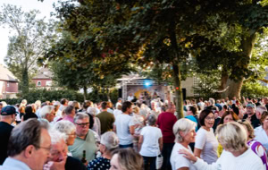 Eine gro&szlig;e Menschenmenge versammelt sich im Freien bei Tageslicht, umgeben von B&auml;umen und Geb&auml;uden.