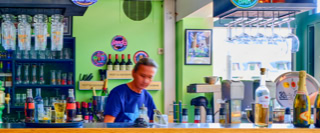 Eine Frau steht hinter einer Bar mit verschiedenen Flaschen und Getr&auml;nken, gr&uuml;ne W&auml;nde im Hintergrund.