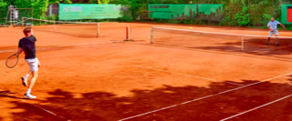 Zwei Personen spielen Tennis auf einem Sandplatz, umgeben von gr&uuml;nen B&auml;umen und Werbebannern.