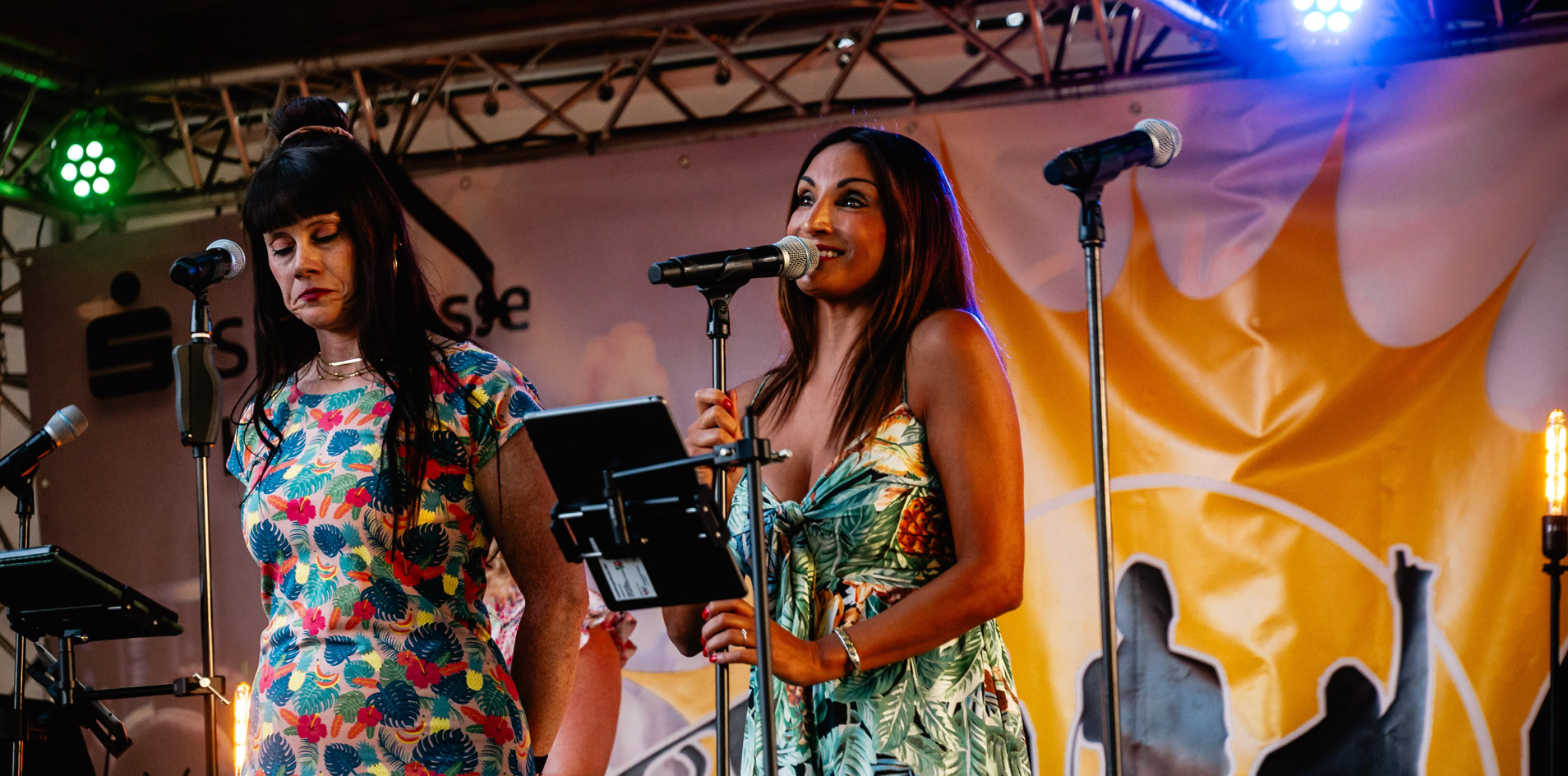 Zwei Frauen singen auf einer B&uuml;hne mit Mikrofonen, umgeben von bunten Lichtern und einem Hintergrund mit Silhouetten.