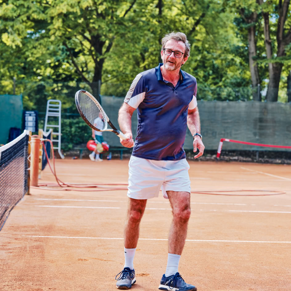 Ein Mann in Sportkleidung steht auf einem Tennisplatz mit einem Tennisschl&auml;ger in der Hand, umgeben von B&auml;umen im Hintergrund.