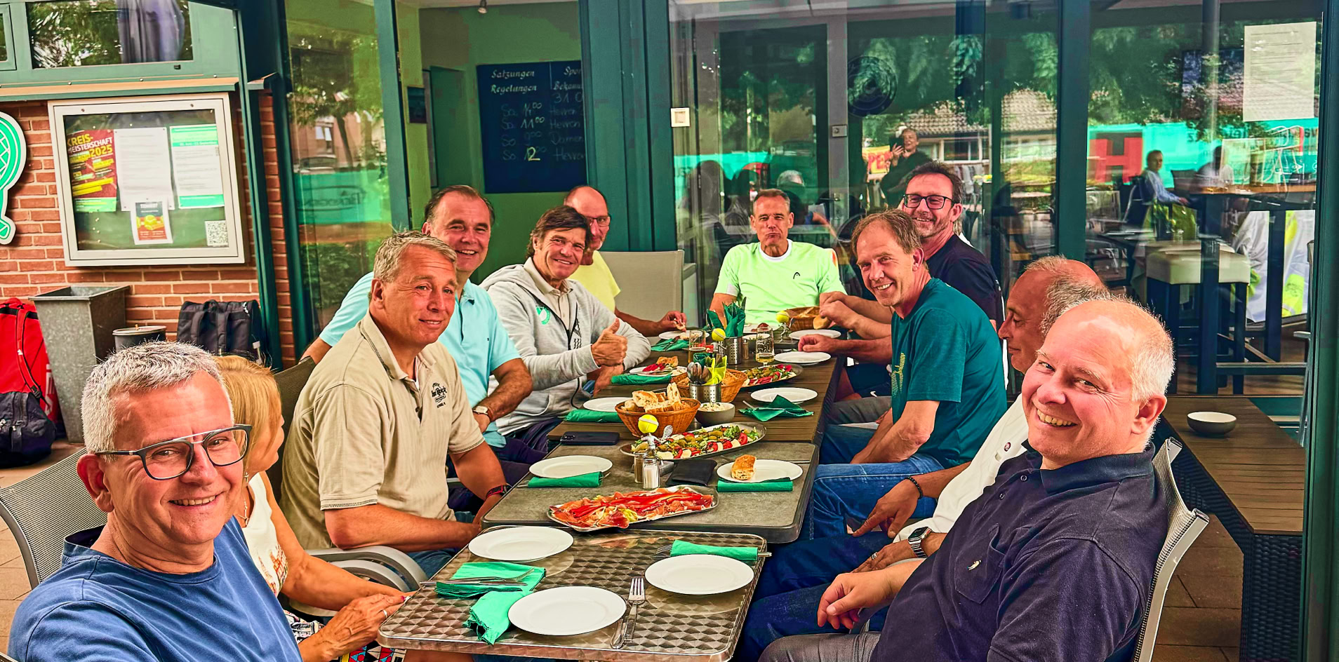 Eine Gruppe von Menschen sitzt um einen Tisch im Freien und l&auml;chelt in die Kamera. Auf dem Tisch stehen Teller und Getr&auml;nke. Im Hintergrund ist ein Geb&auml;ude mit gro&szlig;en Fenstern zu sehen.