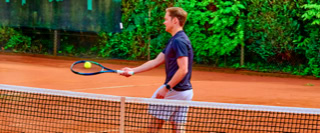 Eine Person spielt Tennis auf einem Sandplatz, umgeben von gr&uuml;ner Vegetation.