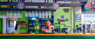 Eine Frau steht hinter einer Bar mit gr&uuml;ner Wand, Gl&auml;sern und Flaschen im Hintergrund.