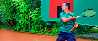 Ein Tennisspieler in gr&uuml;ner Kleidung schl&auml;gt einen Ball auf einem Sandplatz, im Hintergrund sind gr&uuml;ne Pflanzen und ein Schild.