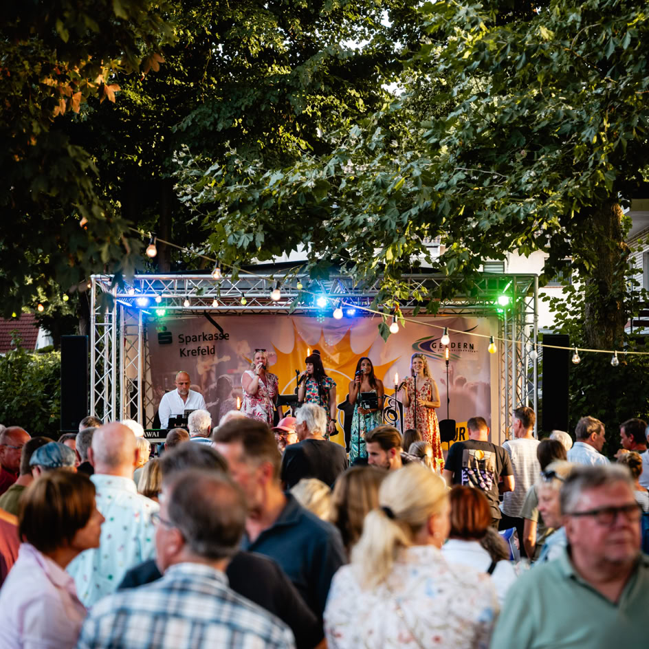Menschenmenge vor einer B&uuml;hne im Freien mit bunten Lichtern, umgeben von B&auml;umen.