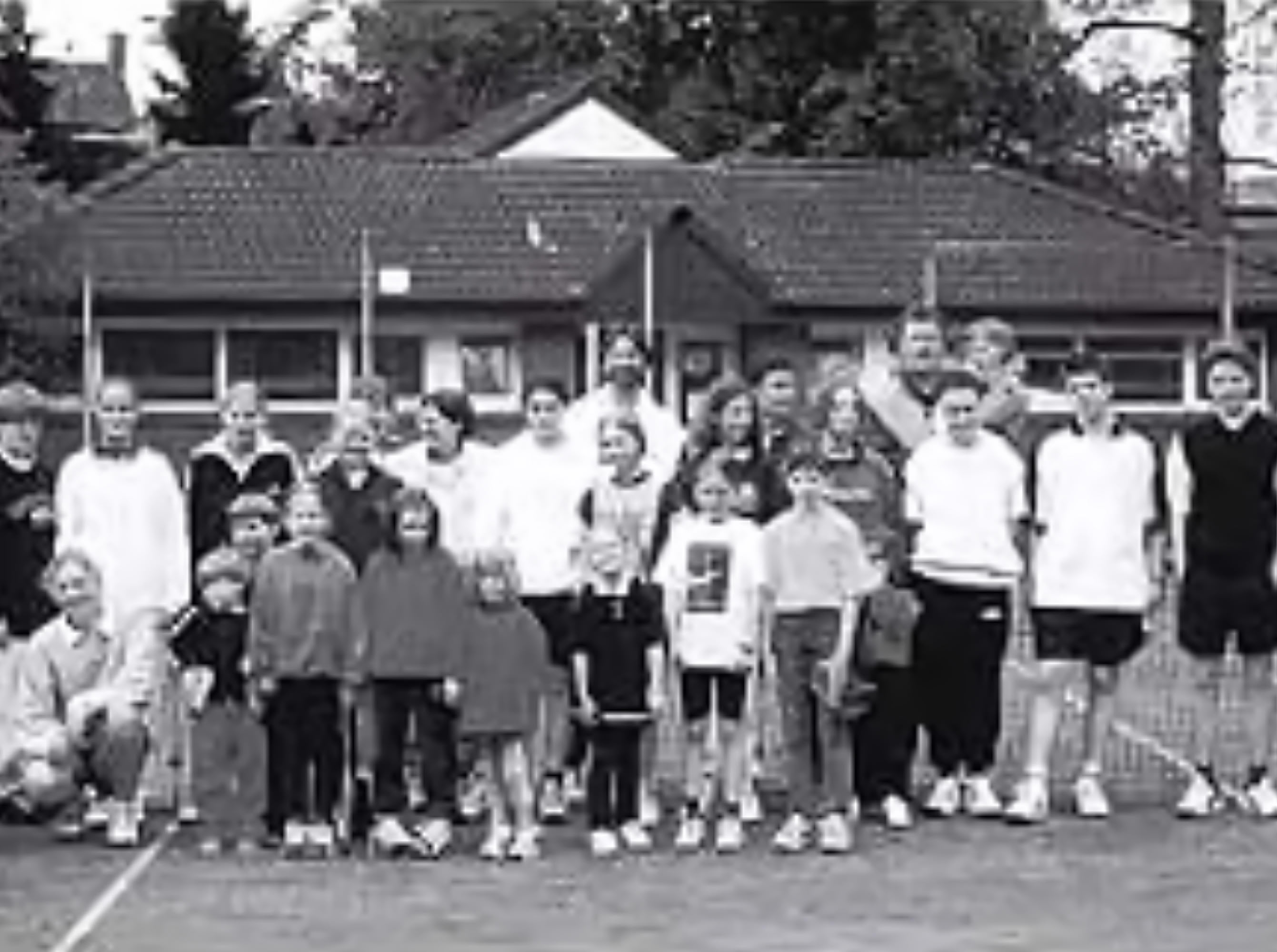 Gruppe von Menschen auf einem Tennisplatz, einige stehen, andere hocken; im Hintergrund sind B&auml;ume und Geb&auml;ude zu sehen.