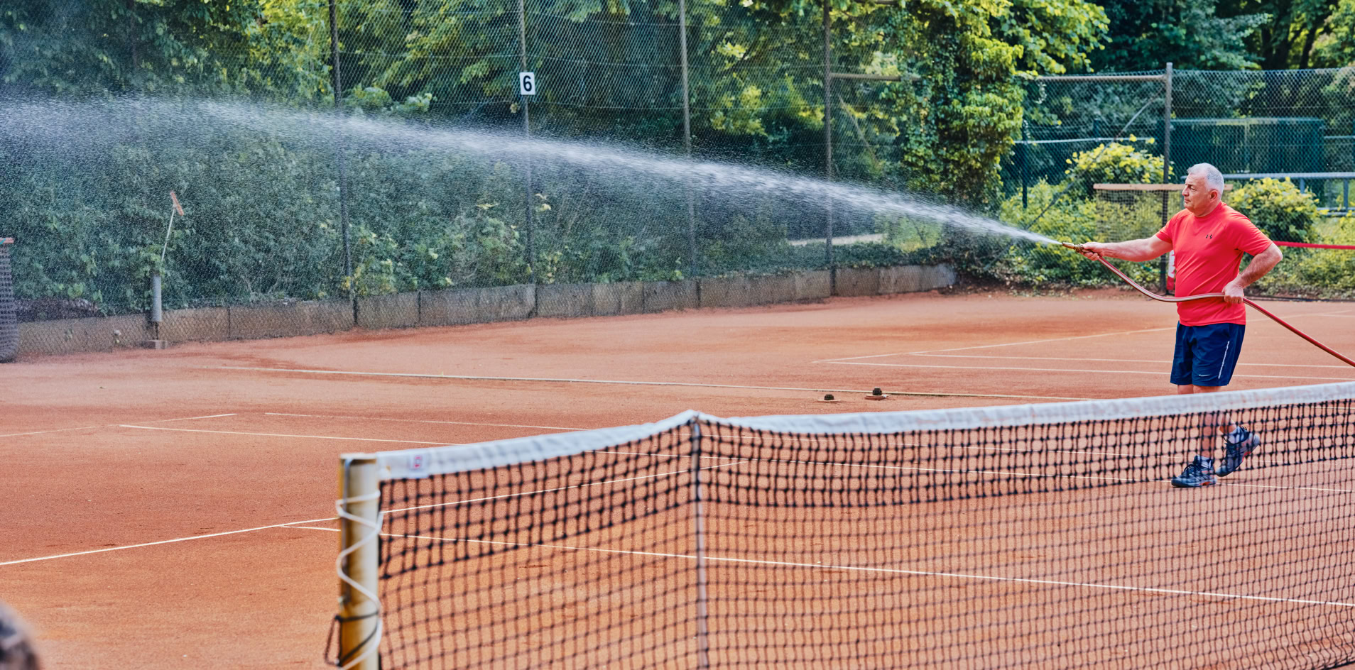 Eine Person bew&auml;ssert einen Sandtennisplatz mit einem Schlauch, umgeben von B&auml;umen und einem Zaun.