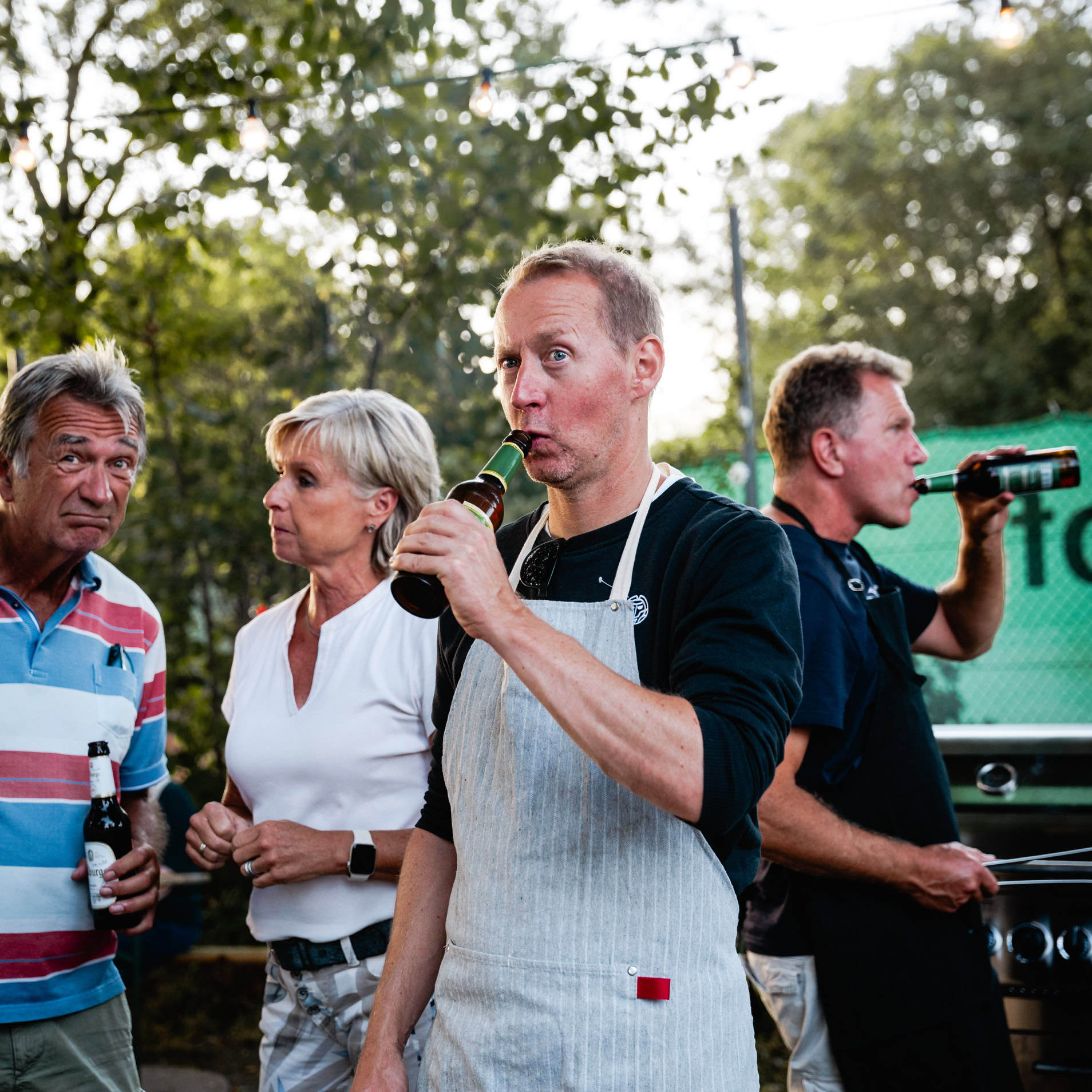 Eine Gruppe von vier Personen steht im Freien, einige halten Flaschen in der Hand. Eine Person tr&auml;gt eine Sch&uuml;rze. Im Hintergrund sind B&auml;ume sichtbar.