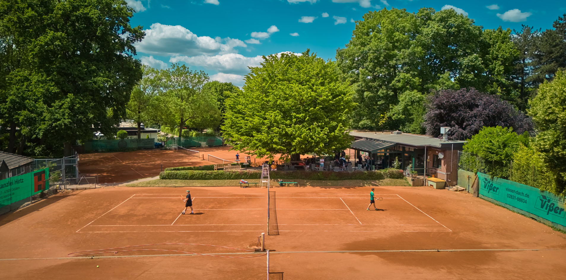 Zwei Personen spielen Tennis auf einem Sandplatz, umgeben von B&auml;umen und einem blauen Himmel mit Wolken.