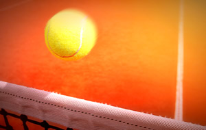 Ein gelber Tennisball fliegt &uuml;ber ein Tennisnetz auf einem Sandplatz.