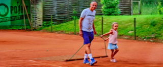 Ein Mann und ein Kind ziehen ein Netz &uuml;ber einen Sandtennisplatz, um den Boden zu gl&auml;tten. Im Hintergrund sind gr&uuml;ne B&auml;ume und ein Zaun zu sehen.