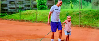 Ein Erwachsener und ein Kind ziehen ein Netz &uuml;ber einen Sandtennisplatz, umgeben von gr&uuml;nem Gras und einem Zaun.