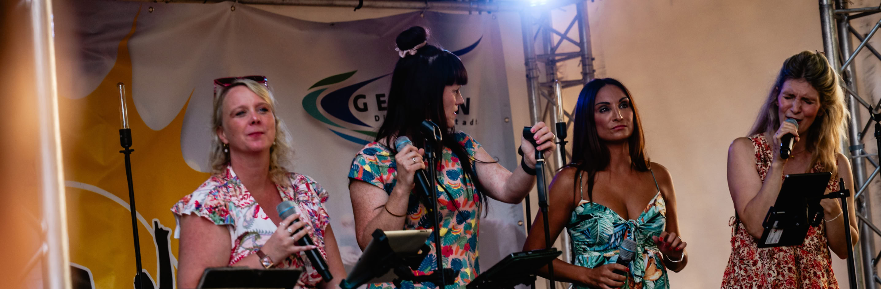 Vier Personen stehen auf einer B&uuml;hne unter einem Metallger&uuml;st mit Lichtern, sie tragen bunte Kleider und halten Mikrofone. Im Vordergrund ist ein Keyboard zu sehen.